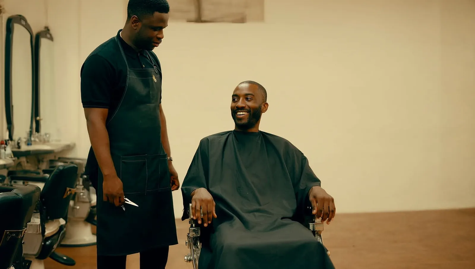 A Black barber in a black apron stands beside a smiling client seated under a cape in a barber chair, the two of them talking.