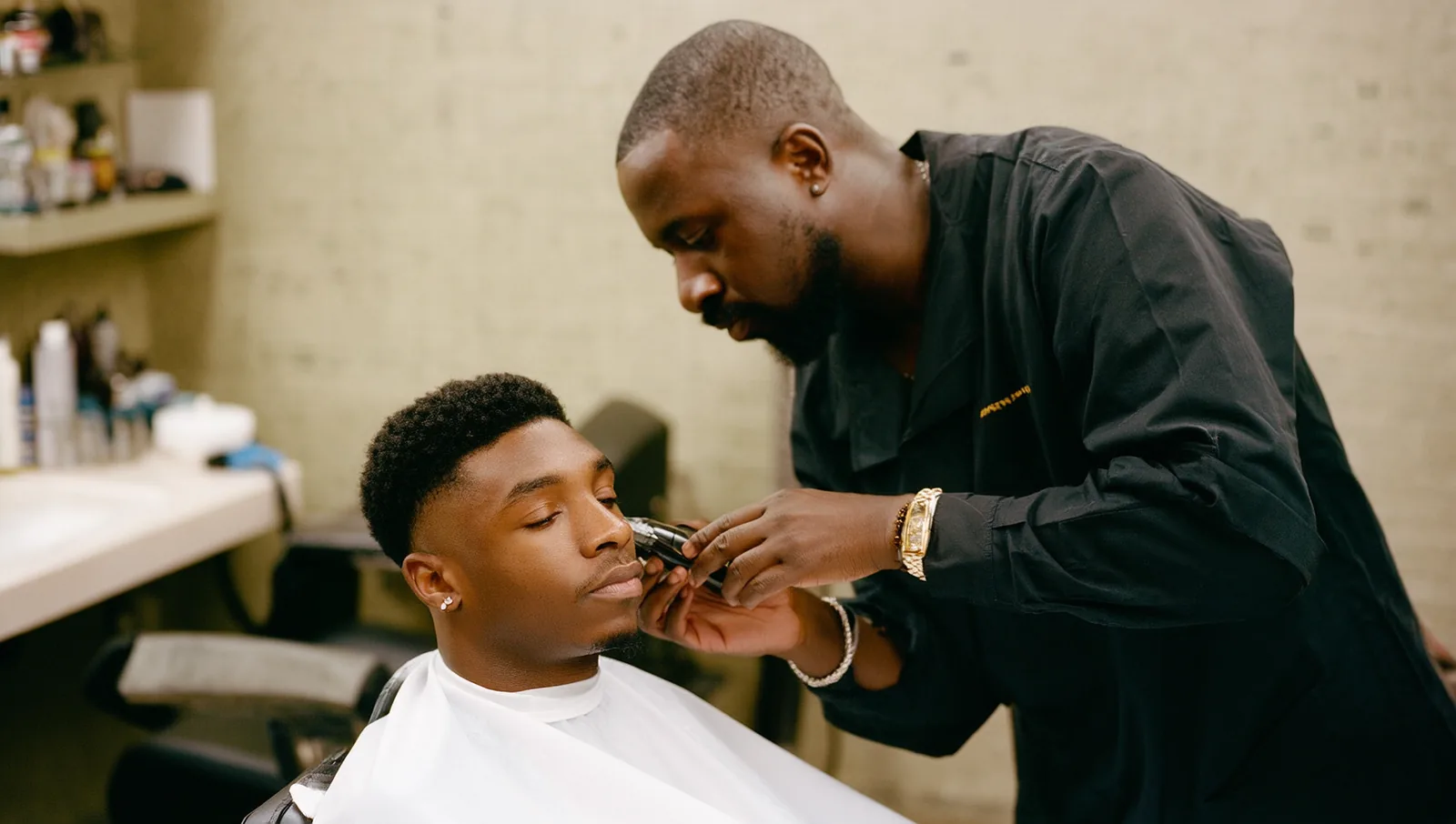 A Black barber details a crisp lineup along a relaxed seated client's hairline at a clean barber station.