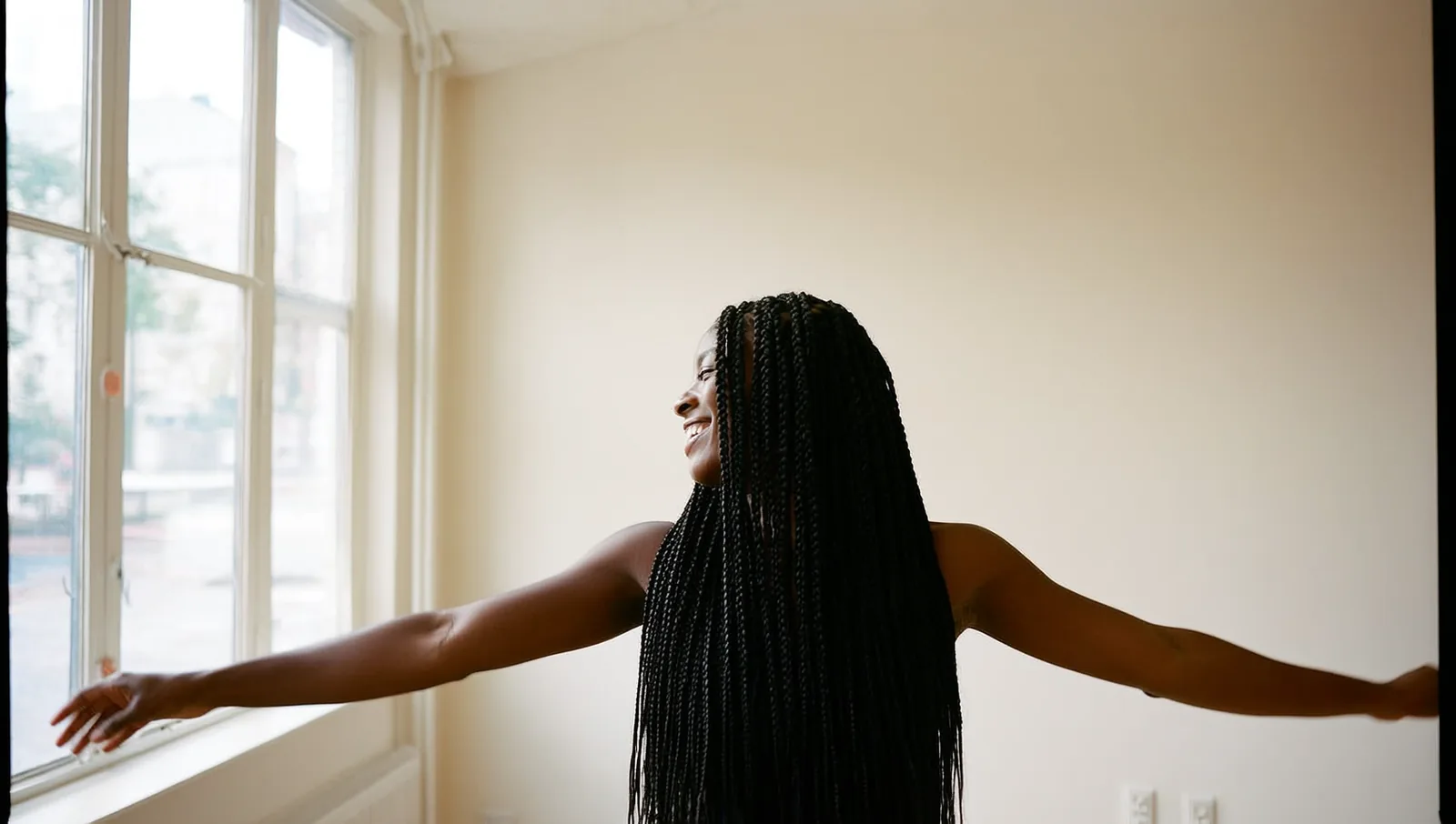 A Black woman with long box braids stands in a bright sunlit room, smiling with her braids falling over one shoulder.