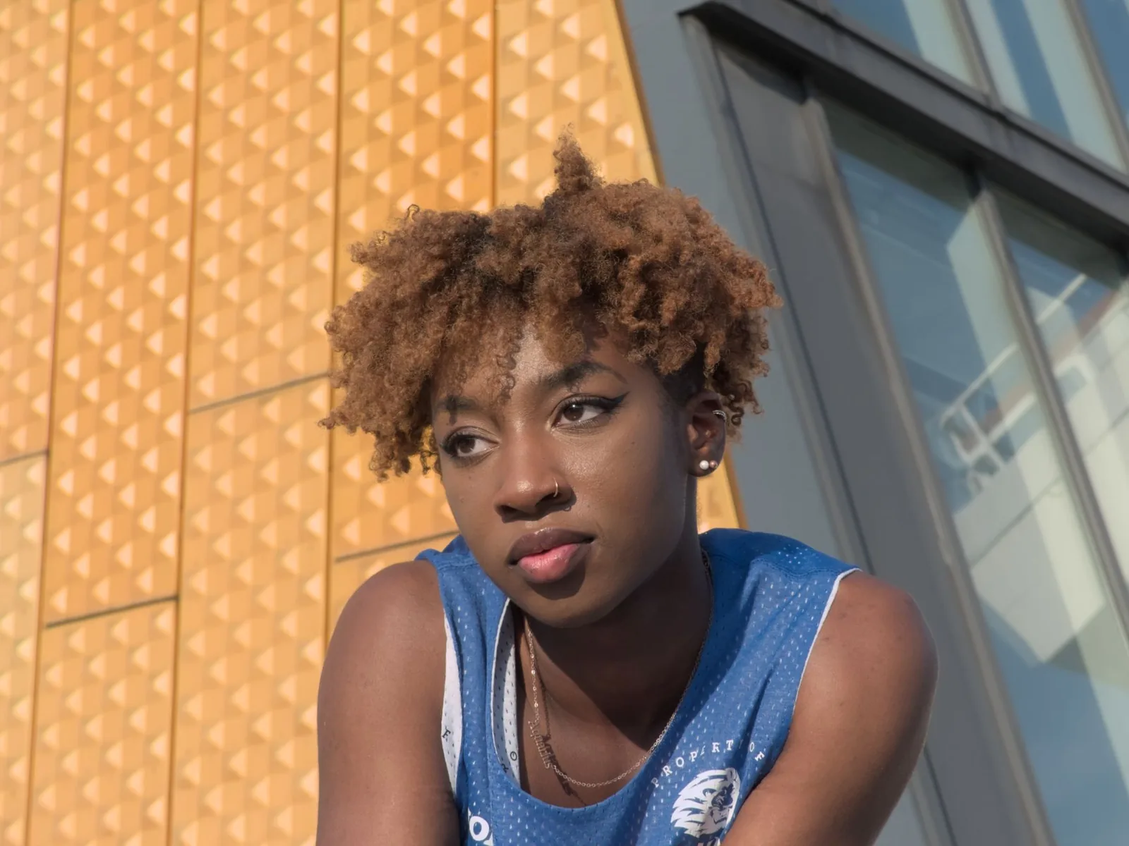 A young woman with a short natural afro sits outdoors in bright daylight beside a gold-panelled building.