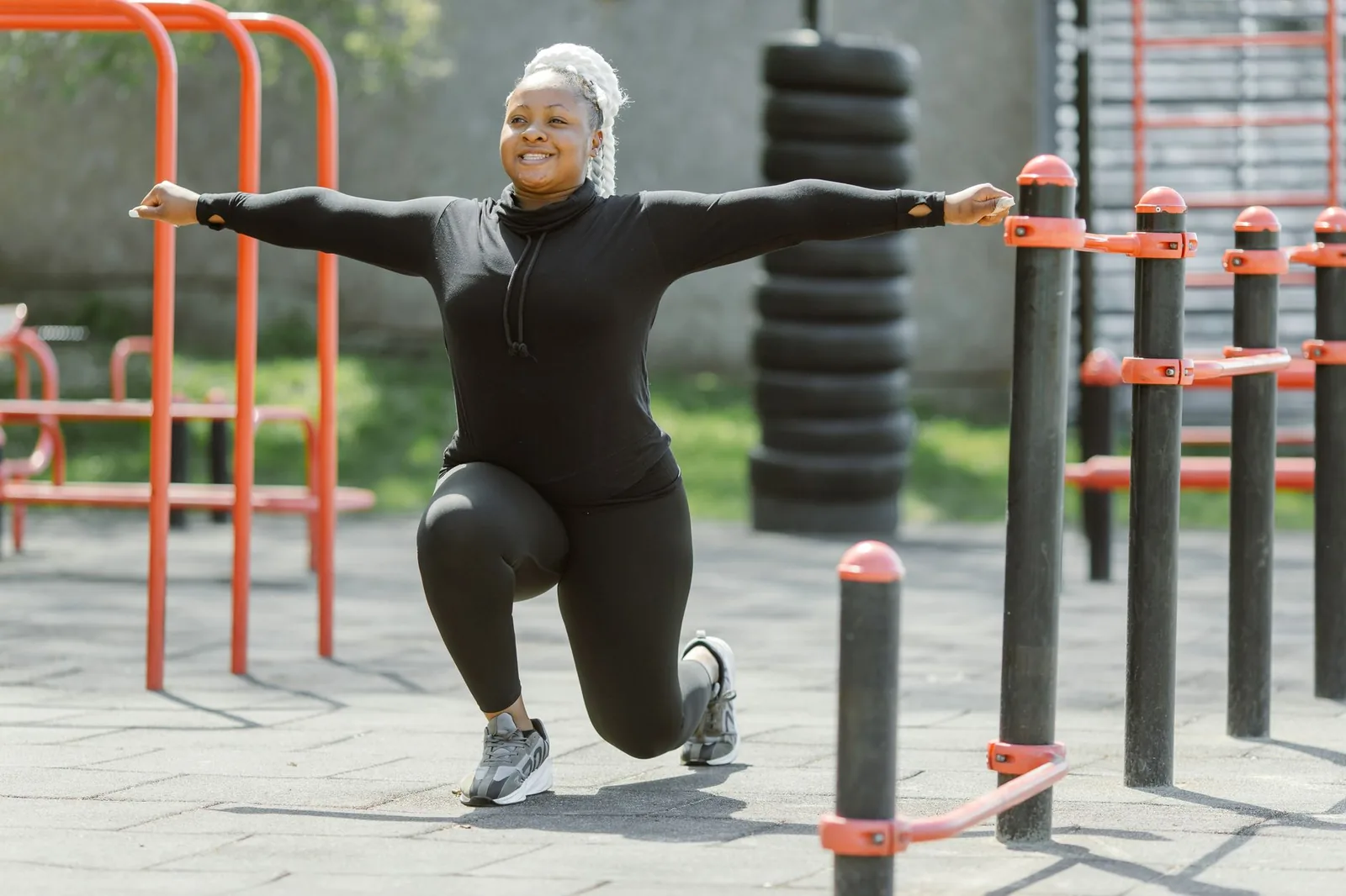 A Black woman in braids and black athletic wear holds a lunge with her arms outstretched at an outdoor calisthenics park.